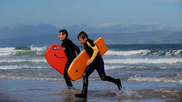 Two young visitors running out of the water holding bodyboards at Portstewart Strand, with the surf visible in the background